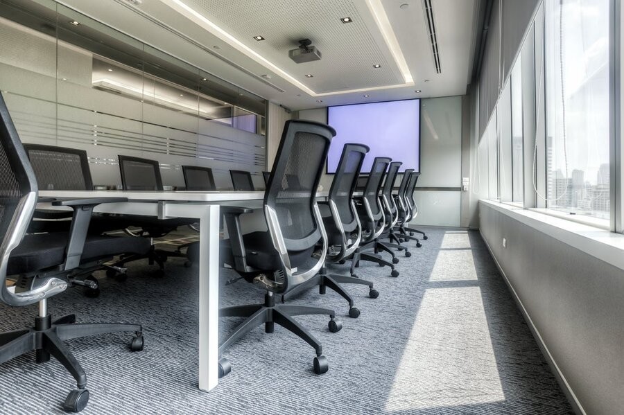 A conference room table in view with windows to the right and a large screen display in the back.